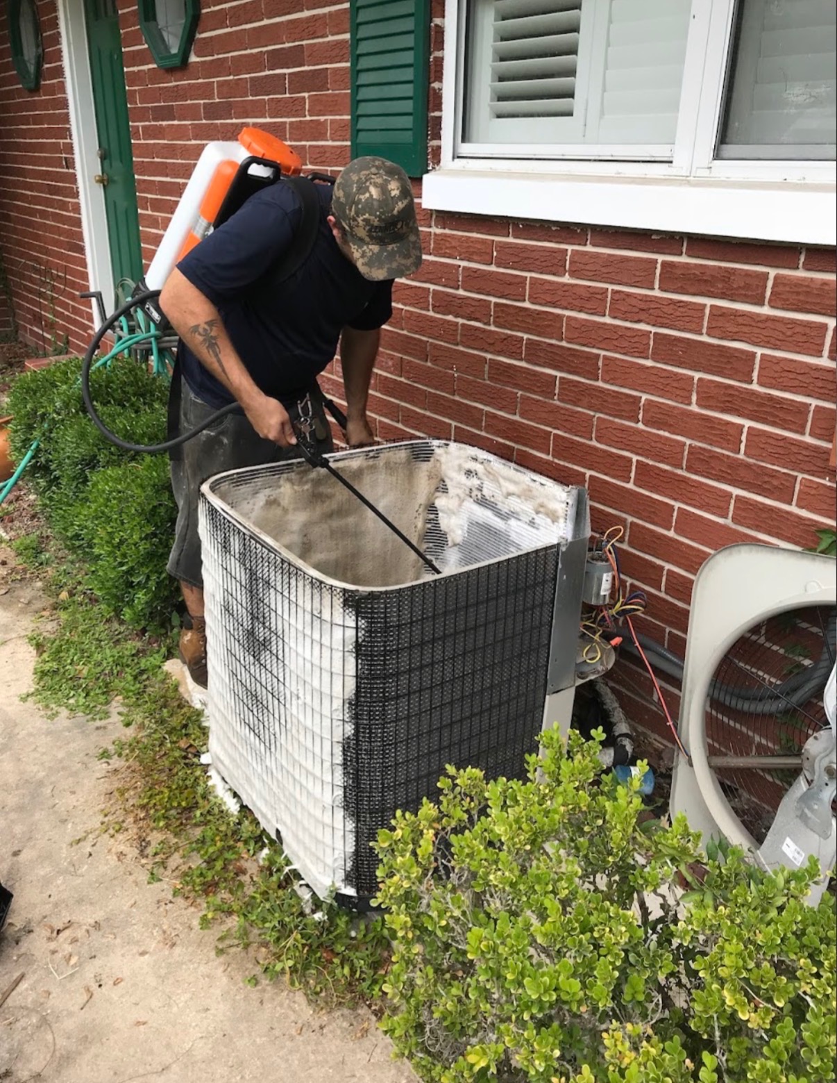 HVAC technician cleaning a dirty condenser coil outside a brick home